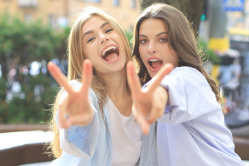 Two young smiling hipster women in summer clothes posing on street.Female showing positive face emotions.