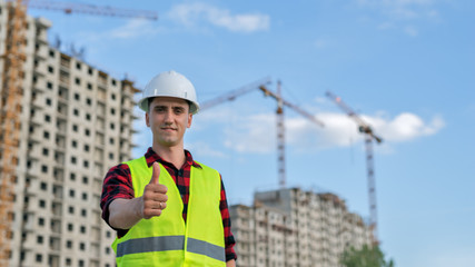 civil engineer in a white helmet on the background of construction, with a raised thumb.