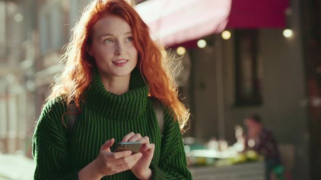 Attractive young ginger woman reading message on smartphone smiling cheerfully walking in street of city center. Portrait of red-headed girl student with mobile phone outdoor.