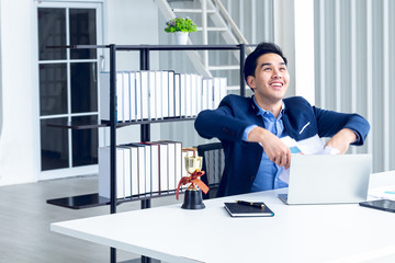 young businessman sitting in a modern office. He has a feel happy and smile about the result of business profits is positive. On his table have a computer laptop tablet pen paper graph.