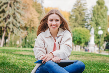 Obraz premium Blonde girl student sitting on the grass and makes notes in a notebook. Student girl smiles and teaches lessons