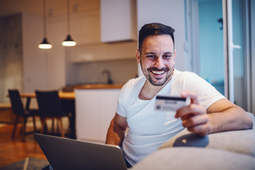 Cheerful caucasian man in pajamas sitting in living room on sofa with laptop in lap and credit card in hand. Morning time. Online shopping concept.