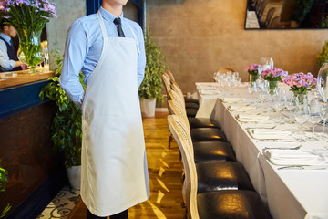 Table setting for guests in a restaurant, on the table are forks and glasses for wine - next to it is a waiter in an apron, flowers in a vase and a candle on the table
