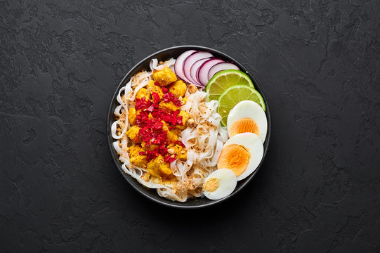 Nan Gyi Thoke in black bowl at dark slate background. Nan gyi Thohk is popular burmese cuisine dish with rice noodles, chicken breasts with spices, eggs, peanuts, lime and red onion. Top view