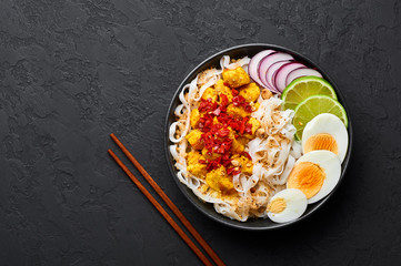 Nan Gyi Thoke in black bowl at dark slate background. Nan gyi Thohk is popular burmese cuisine dish with rice noodles, chicken breasts with spices, eggs, peanuts, lime and red onion. Top view