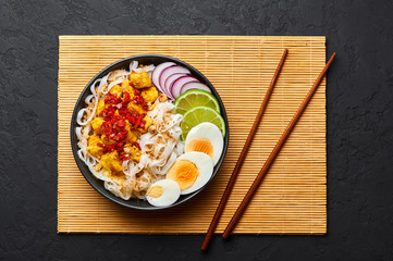 Nan Gyi Thoke in black bowl at dark slate background. Nan gyi Thohk is popular burmese cuisine dish with rice noodles, chicken breasts with spices, eggs, peanuts, lime and red onion. Top view