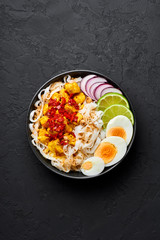 Nan Gyi Thoke in black bowl at dark slate background. Nan gyi Thohk is popular burmese cuisine dish with rice noodles, chicken breasts with spices, eggs, peanuts, lime and red onion. Top view
