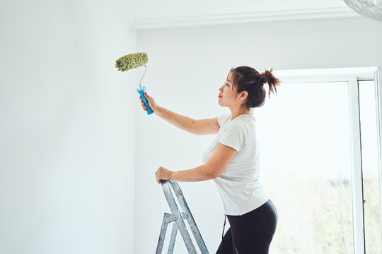 Repair in the apartment, the girl makes repairs in the apartment, paint the walls with a roller, standing on a ladder