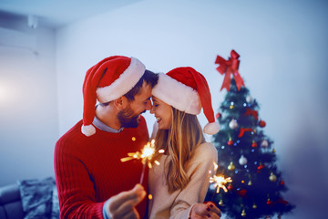 Obraz premium Happy caucasian couple with santa hats on heads holding sprinklers while cuddling and standing in living room. In background is christmas tree.
