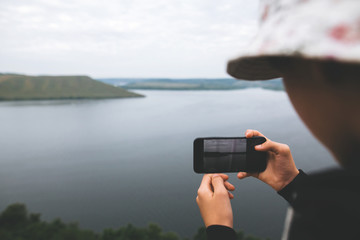 Hipster teenager taking photo on phone of amazing view on river while standing on top of rock mountain. Young stylish guy exploring and traveling. Atmospheric moment. Copy space