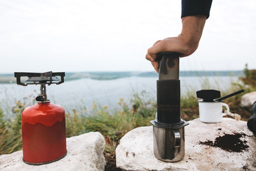 Traveler pressing hot water through coffee in aeropress on cliff at lake, brewing alternative...
