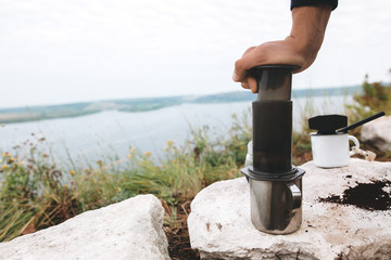 Traveler pressing aeropress on metal mug on cliff at lake, brewing alternative coffee at camping....