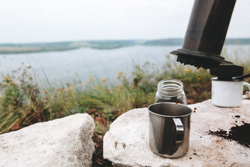 Obraz premium Coffee drops from aeropress on metal mug on cliff at lake, brewing alternative coffee at camping. Making hot drink at picnic outdoors. Trekking and hiking in mountains