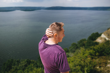 Fototapeta na wymiar Stylish hipster traveler in purple shirt standing on top of rock mountain with amazing view on river. Young brutal guy exploring and traveling, back view. Copy space