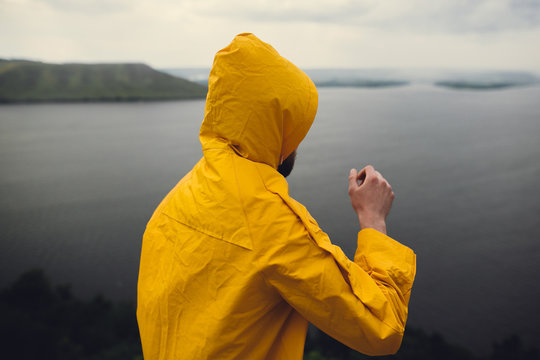 Hipster traveler in yellow raincoat standing on cliff and looking at lake in windy moody day. Wanderlust and travel concept. Man hiking in Norway on foggy day. Atmospheric moment