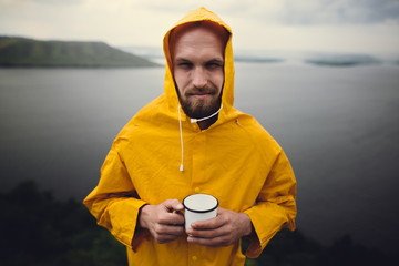 Hipster traveler in yellow raincoat holding metal mug and standing on cliff in rainy windy day with...
