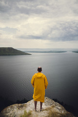 Fototapeta na wymiar Traveler in yellow raincoat standing on cliff and looking at lake in rainy windy day. Wanderlust and travel concept. Hipster man hiking in Norway on foggy day. Atmospheric moment
