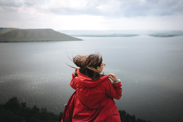 Traveler girl in red raincoat and with windy hair standing on top of rock mountain with beautiful...