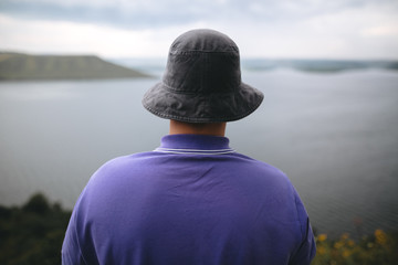 Hipster traveler in purple shirt  and bucket hat standing on top of rock mountain with amazing view on river. Young camper guy exploring and traveling, back view. Copy space