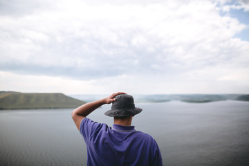 Hipster traveler in purple shirt  and bucket hat standing on top of rock mountain with amazing view on river. Young camper guy exploring and traveling, back view. Copy space - obrazy, fototapety, plakaty