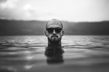 Brutal bearded man in sunglasses emerge in lake waves. Man head above water in lake in rainy foggy...
