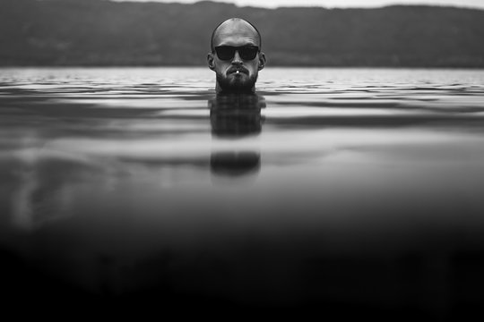 Brutal Bearded Man With Cigarette And In Sunglasses Emerge In Lake. Man Head Smoking Above Water In Lake In Rainy Foggy Day, Atmospheric Moment. Wanderlust. Creative Black And White Photo