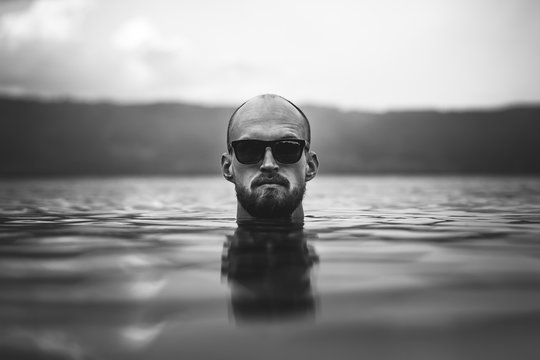 Brutal bearded man in sunglasses emerge in lake waves. Man head above water in lake in rainy foggy day, atmospheric moment. Wanderlust. Creative black and white photo
