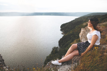 Traveler hipster girl in hat and windy hair sitting on top of rock mountain with wildflowers,...