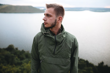 Hipster traveler standing on top of rock mountain and enjoying amazing view on river. Stylish guy in green windbreaker exploring and traveling. Atmospheric tranquil moment. Copy space © sonyachny