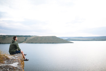 Fototapeta na wymiar Hipster traveler sitting on top of rock mountain and enjoying amazing view on river. Stylish guy exploring and traveling. Atmospheric tranquil moment. Copy space