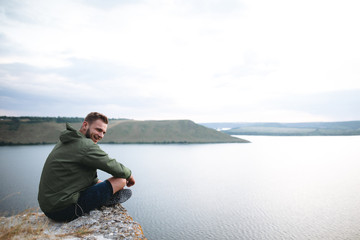 Hipster traveler sitting on top of rock mountain and enjoying amazing view on river. Stylish guy in green windbreaker exploring and traveling. Atmospheric tranquil moment. Copy space © sonyachny