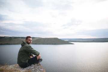 Hipster traveler sitting on top of rock mountain and enjoying amazing view on river. Stylish guy exploring and traveling. Atmospheric tranquil moment. Copy space