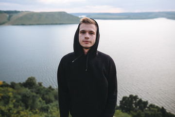 Fototapeta premium Portrait of hipster traveler in windbreaker on top of rock mountain with amazing view on river. Stylish guy exploring and traveling. Atmospheric moment. Copy space