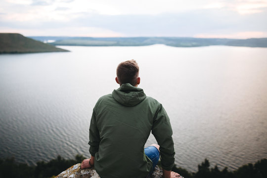 Hipster traveler sitting on top of rock mountain and enjoying amazing view on river. Stylish guy in green windbreaker exploring and traveling. Atmospheric tranquil moment. Copy space
