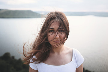 Traveler girl with windy hair relaxing on top of rock mountain, enjoying beautiful sunset view on river. Atmospheric moment. Copy space. Calm portrait of brunette woman in white shirt