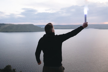 Protest concept. Brutal man holding blue flare torch in hand, standing on top of hill in evening...