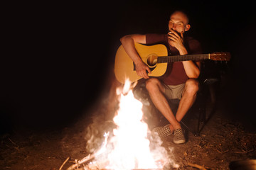 Hipster man smoking cigarette and holding acoustic guitar at big bonfire at camp, chilling at camp...