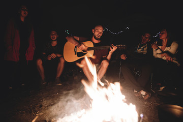 Hipster man playing on acoustic guitar and singing song with friends travelers at big bonfire at...