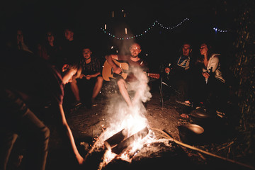 Hipster man playing on acoustic guitar and singing song with friends travelers at big bonfire at...