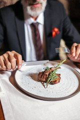 Close up of tasty looking piece of baked hot lamb meat prepared with sauce and prettified with greenery served on big round plate. Male hands ready to cut delicious meat for pieces.