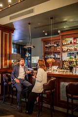 Self confident attractive man in formal suit with unbuttoned jacket, holds delicious wine, looking straight at long haired brunette sits on leather chair, young couple spending time in restaurant
