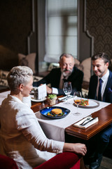 Group of generous friends enjoy dinner in beautiful restaurant, close up of blonde woman sitting with back to camera at table with delicious meal, right side shot