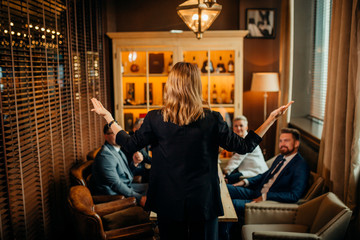 Friendly longhaired business lady in black suit standing in front of active colleagues with her back to camera, stretching arms and explaining lie of matters at cozy small study