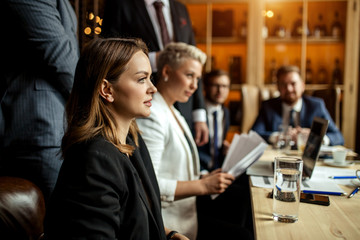 charming brunette woman with long hair in black formal suit sitting at light coloured table at conference listening attentively the speaker not looking at camera