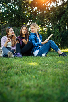 Three friends spend their free time sitting in the park on green lawn. Young pretty girls talking, smiling, looking photo at smartphone. Concept of female friendship. Vertical image.