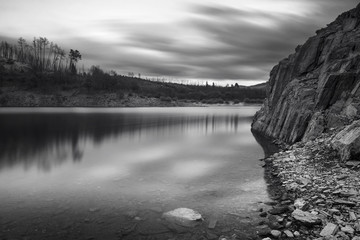 Riverscape on a cloudy early evening with water and rocks on foreground and a beach and trees on a hill in the background, Black and White Photo, Fronhas dam reservoir, Alva river, Fronhas, Portugal