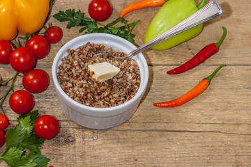 Cooked buckwheat porridge in a deep plate with butter on a wooden background with vegetables.