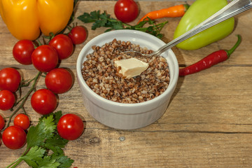 Cooked buckwheat porridge in a deep plate with spoon on a wooden background with vegetables. Copy space