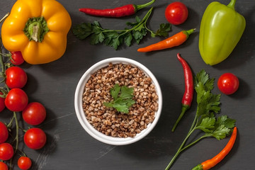 Cooked buckwheat porridge in a deep plate on a gray wooden background with vegetables. Top view