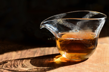 Close-up of a Chinese glass teapot with black fresh tea standing on a table against a dark background with the sunlight shining through it